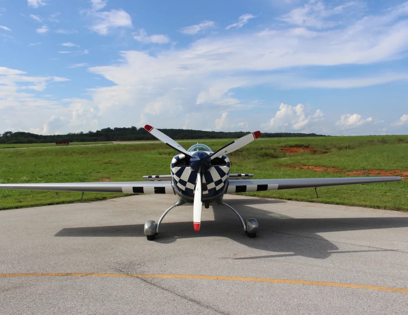 The N51E Extra EA-300/L aircraft is seen from the front, parked on an airfield with green fields in the background.
