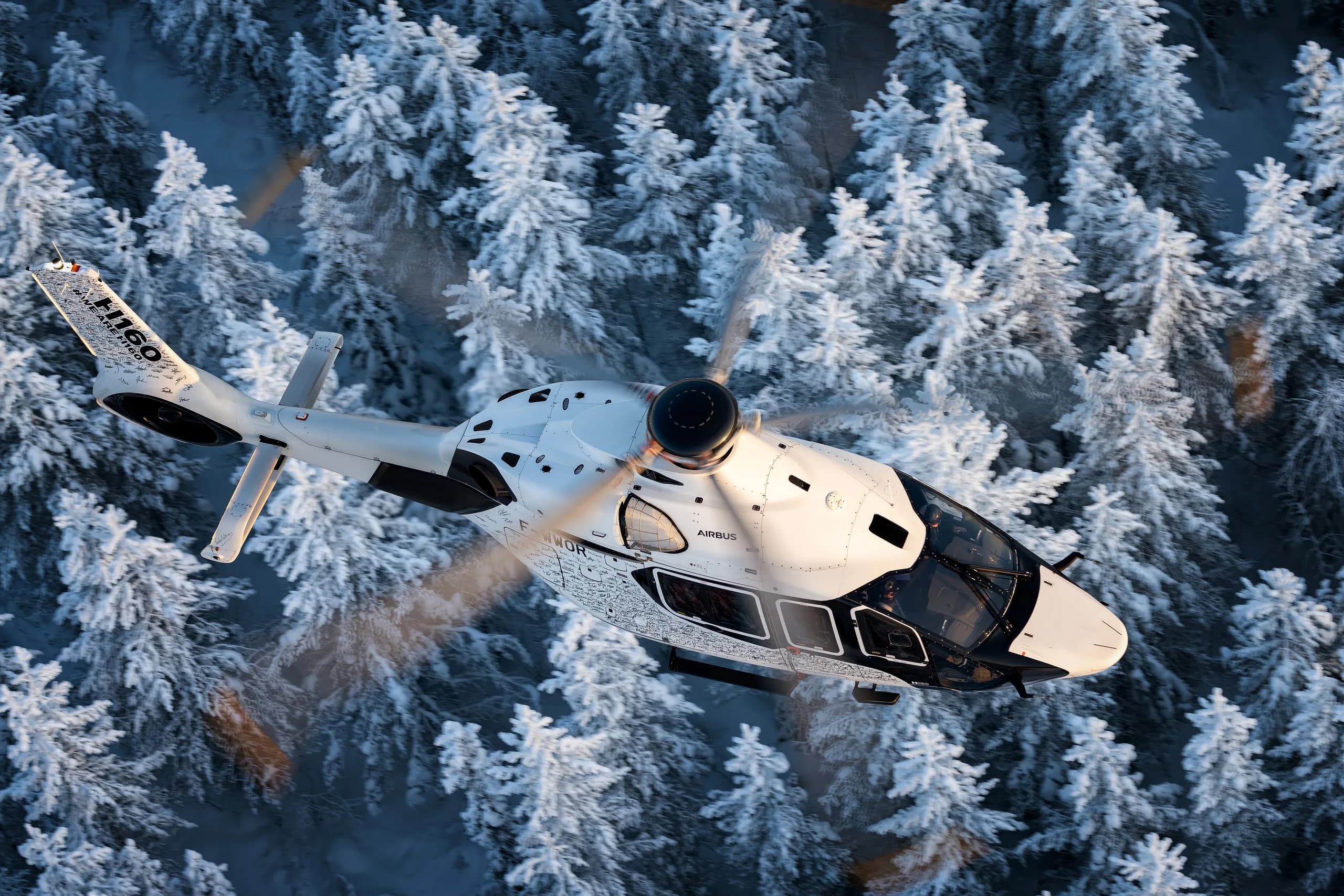 An Airbus H160 helicopter flying over a snowy forest.