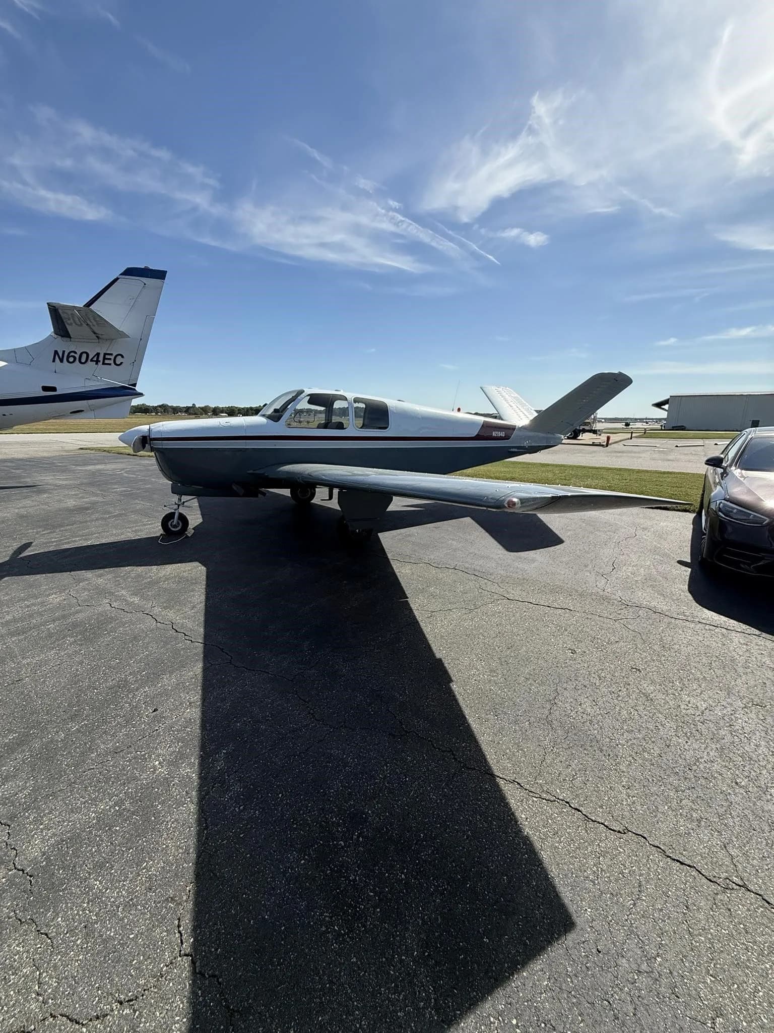 Side view of the N2194D Beechcraft Bonanza V-tail aircraft, with a white and grey paint scheme and red stripe, parked on an airfield next to another aircraft.
