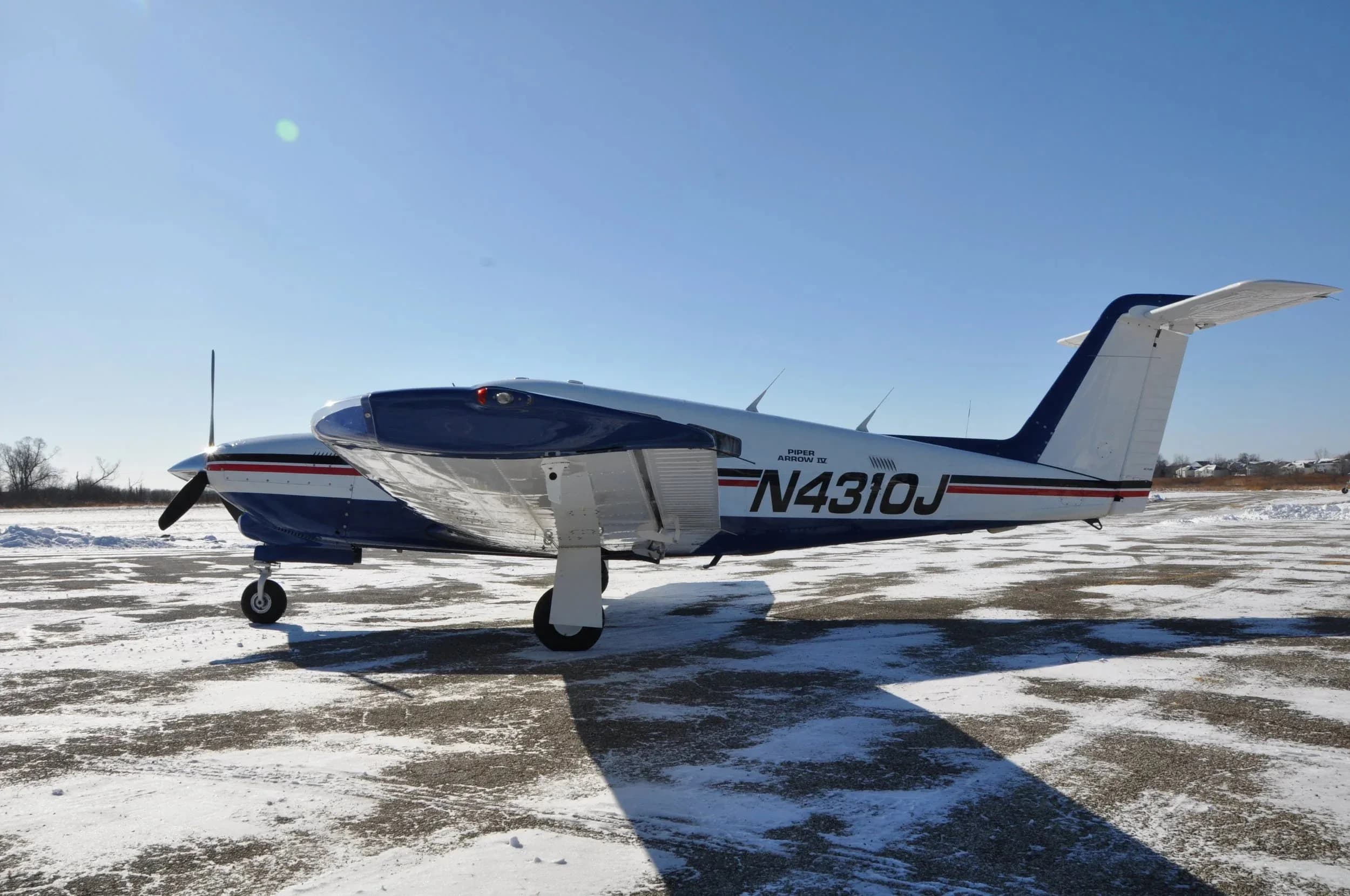 A full side view of the white and blue Piper Arrow IV aircraft with registration N4310J parked on a snowy tarmac, showing the underside of the wing.