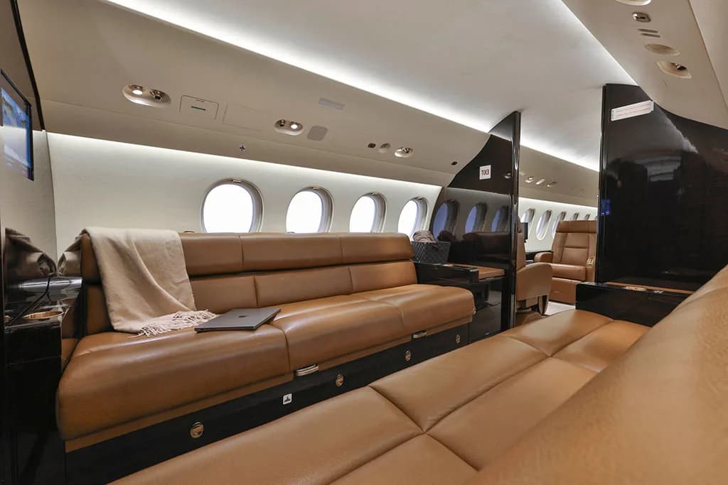 A section of the business jet's cabin features a long tan leather sofa with a blanket and laptop, facing large oval windows.