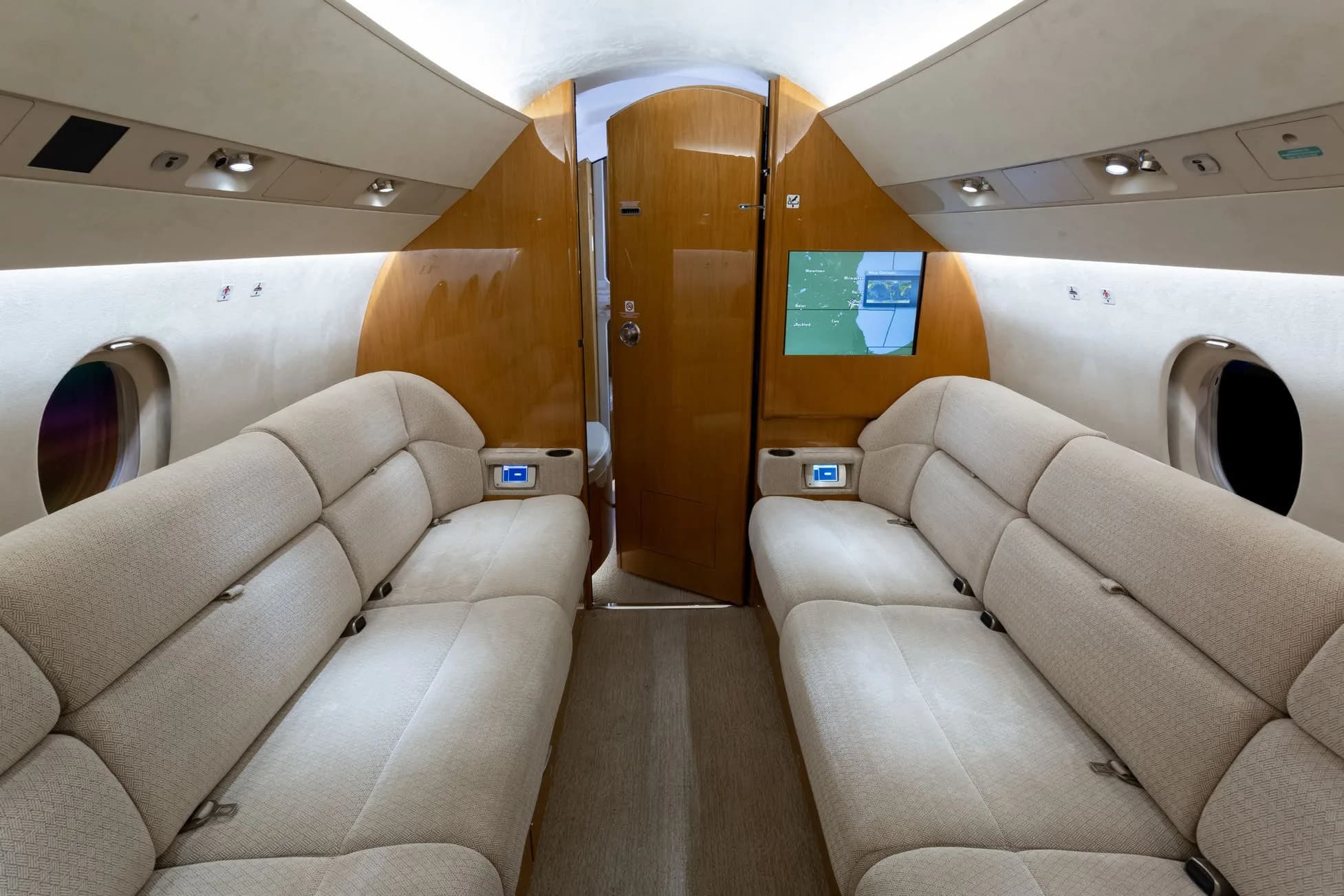 A wide-angle view of the private jet cabin, showing two opposing beige fabric sofas and a screen displaying a map at the far end.