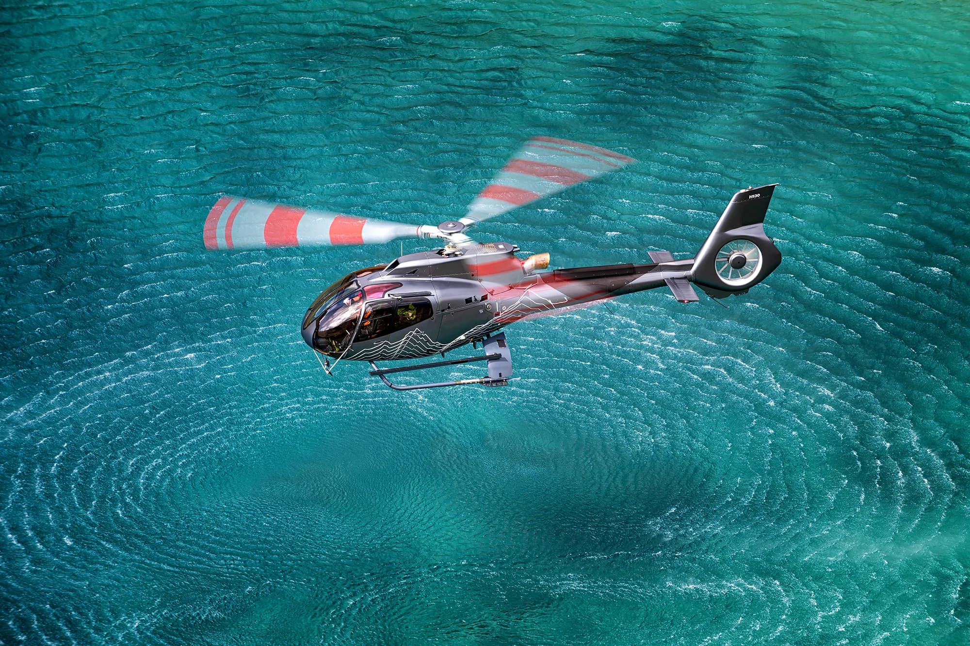 A grey and red Airbus H130 helicopter flies over clear turquoise water, with its main rotor blades blurred in motion.
