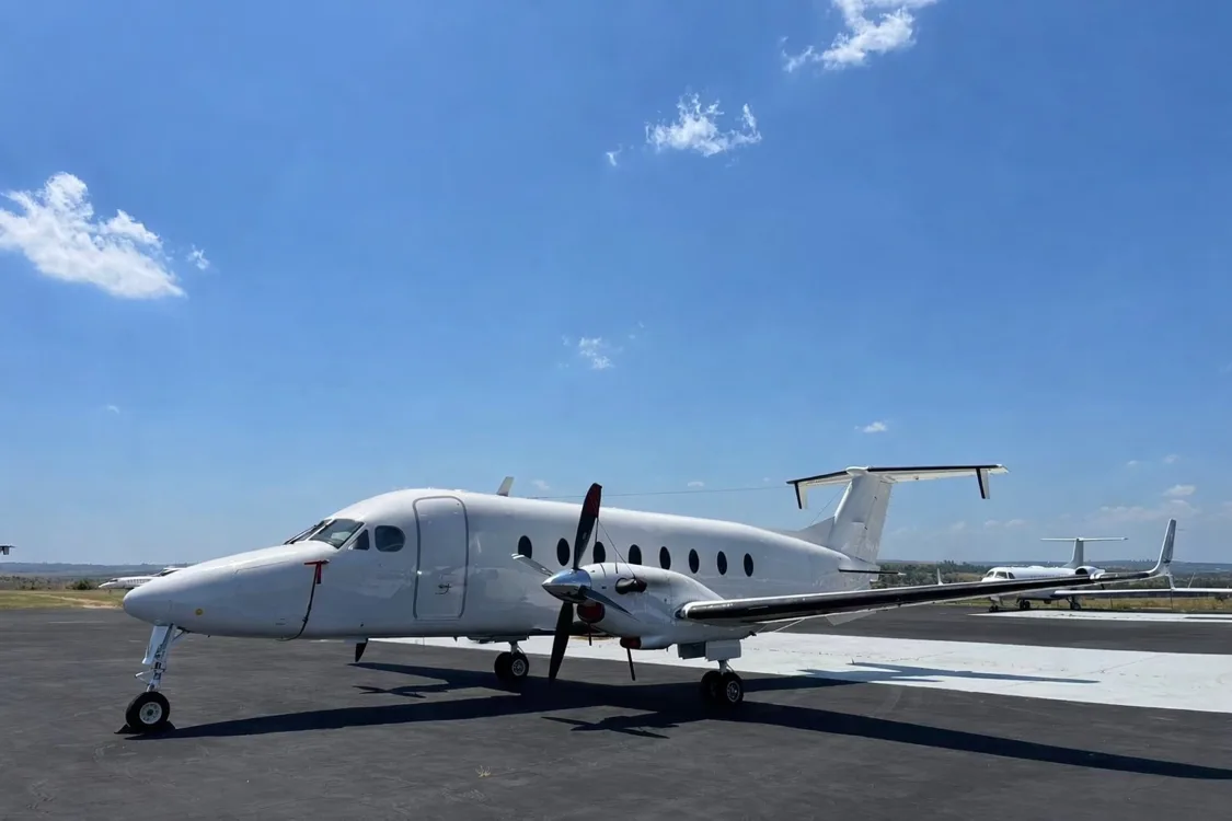A white twin-turboprop aircraft with red propeller tips parked on an asphalt tarmac, viewed from the front-left.