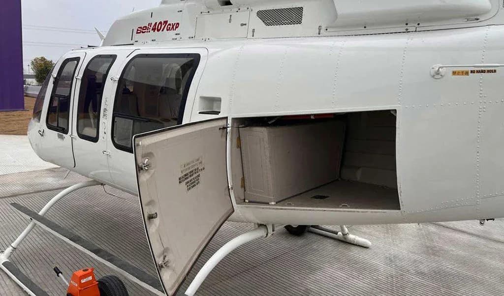 A close-up side view of a white Bell 407GXP helicopter with its cargo compartment door open, revealing an empty storage space.