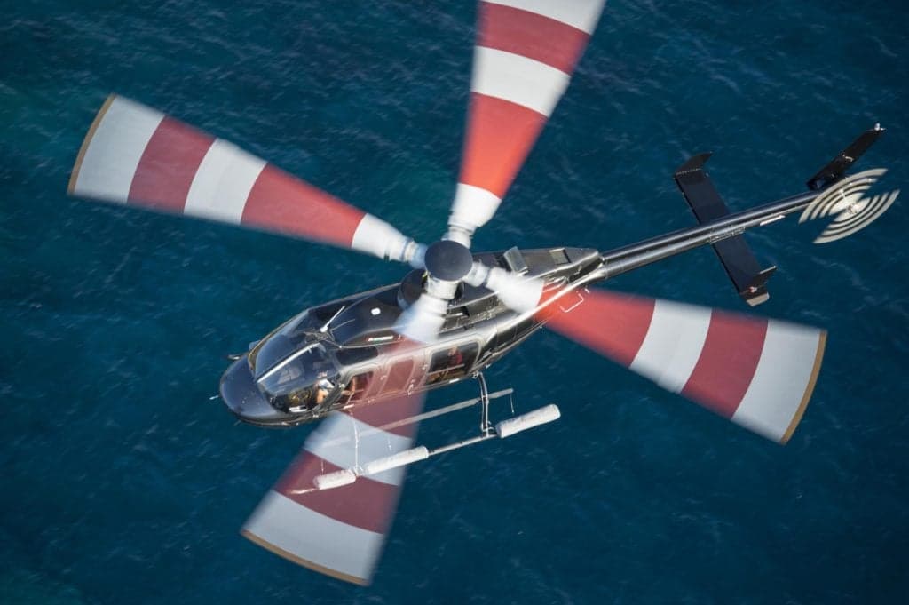 Overhead view of a Bell 407GXi helicopter with red and white striped rotor blades over the ocean.