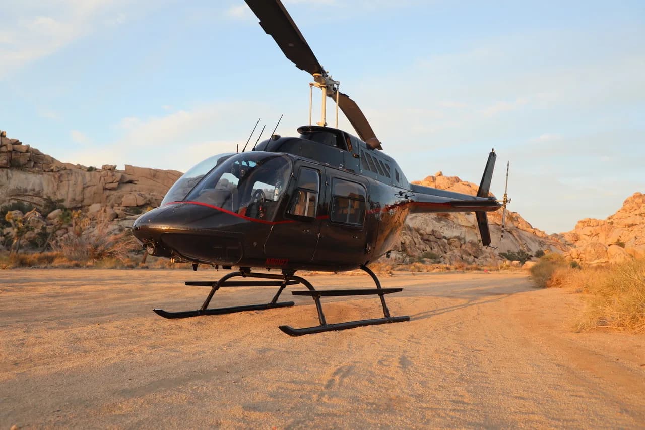 A black Bell 206 JetRanger helicopter with red accents sits on a dirt landing pad in a rocky desert landscape at sunset.