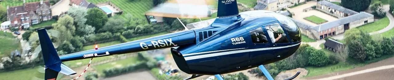 A blue Robinson R66 helicopter in flight over a rural landscape.
