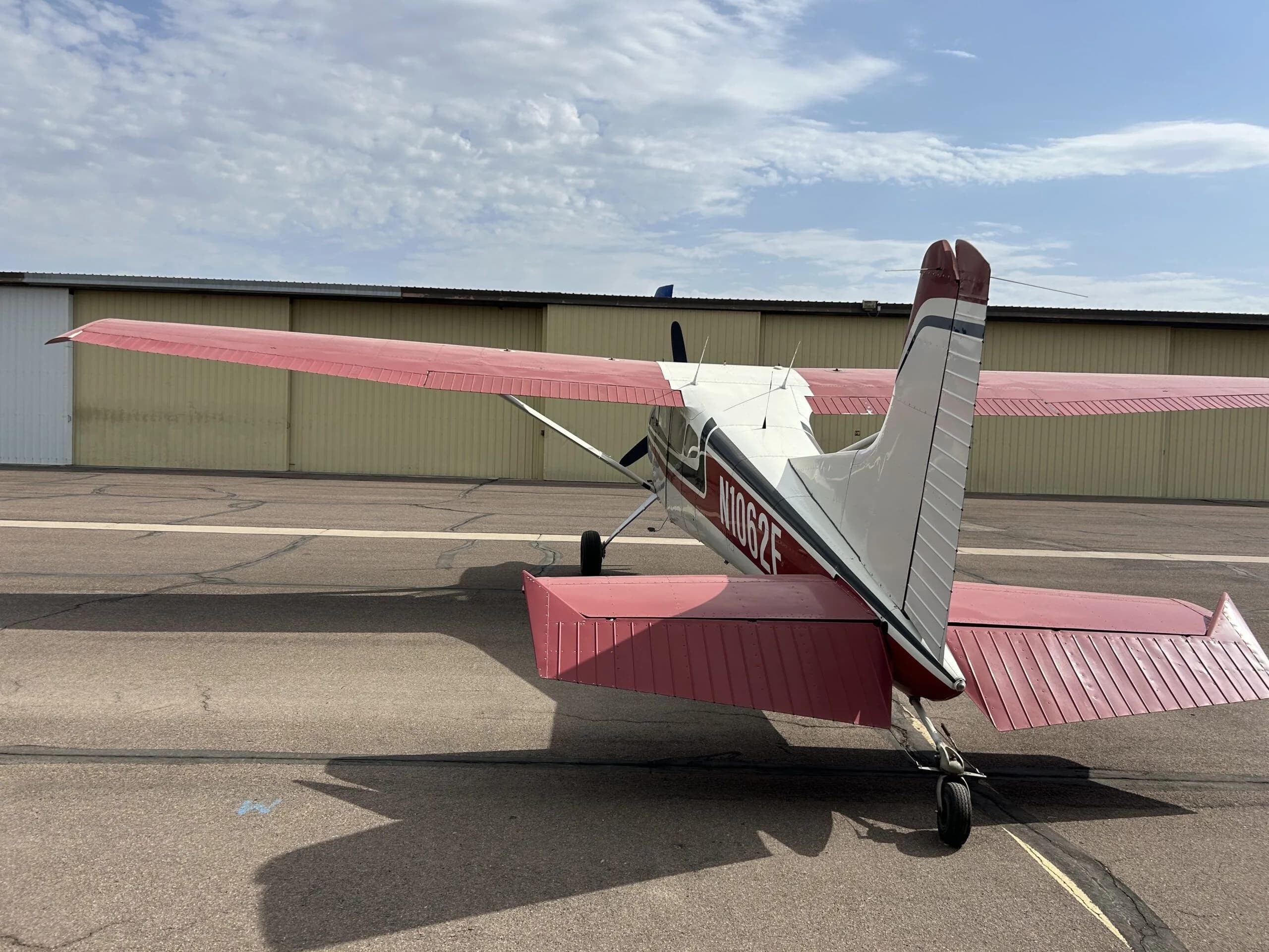 A white, black, and red Cessna 180 aircraft (N1062F) parked on an airport tarmac, viewed from directly behind.