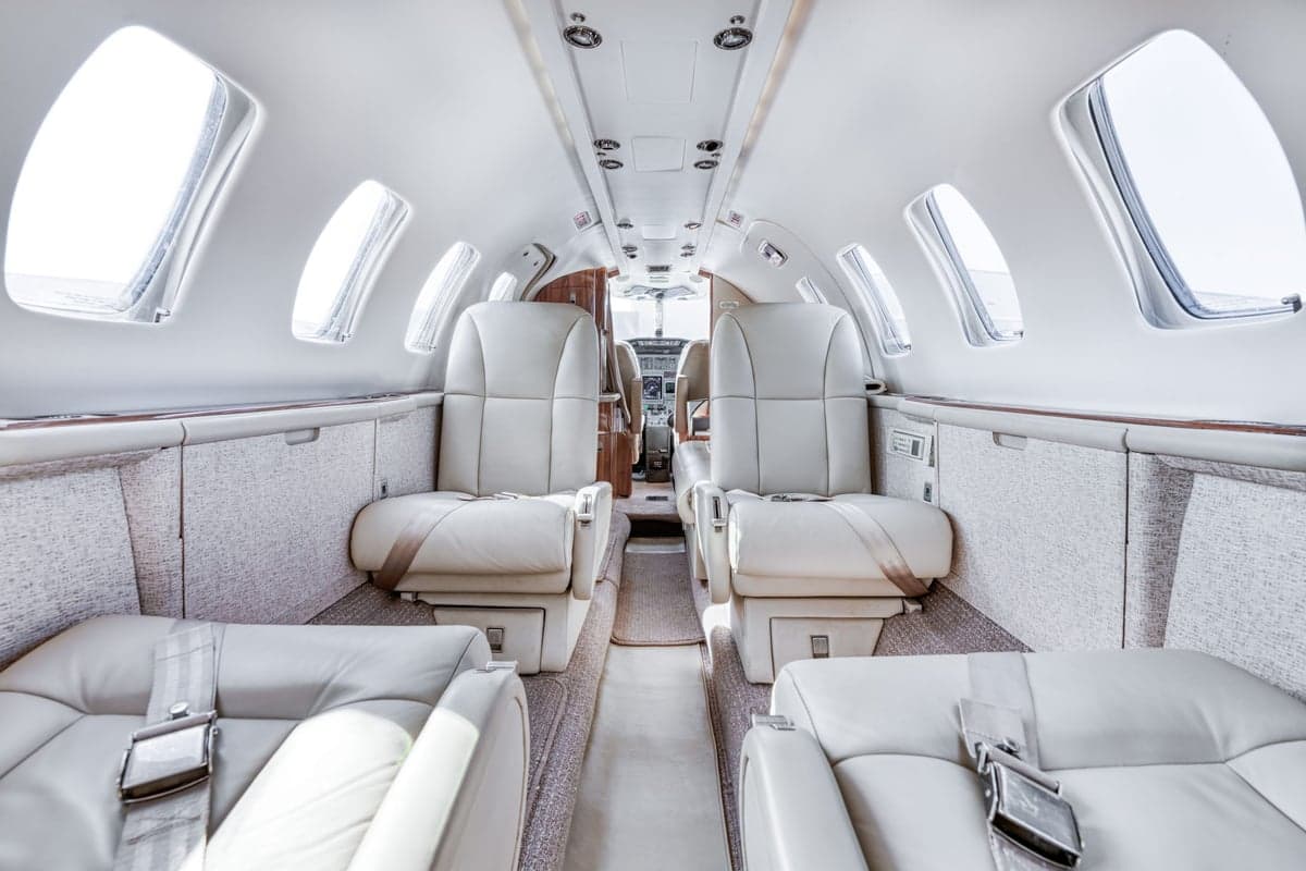 The luxurious interior of a private jet features light cream leather seats and a narrow aisle leading towards the cockpit.