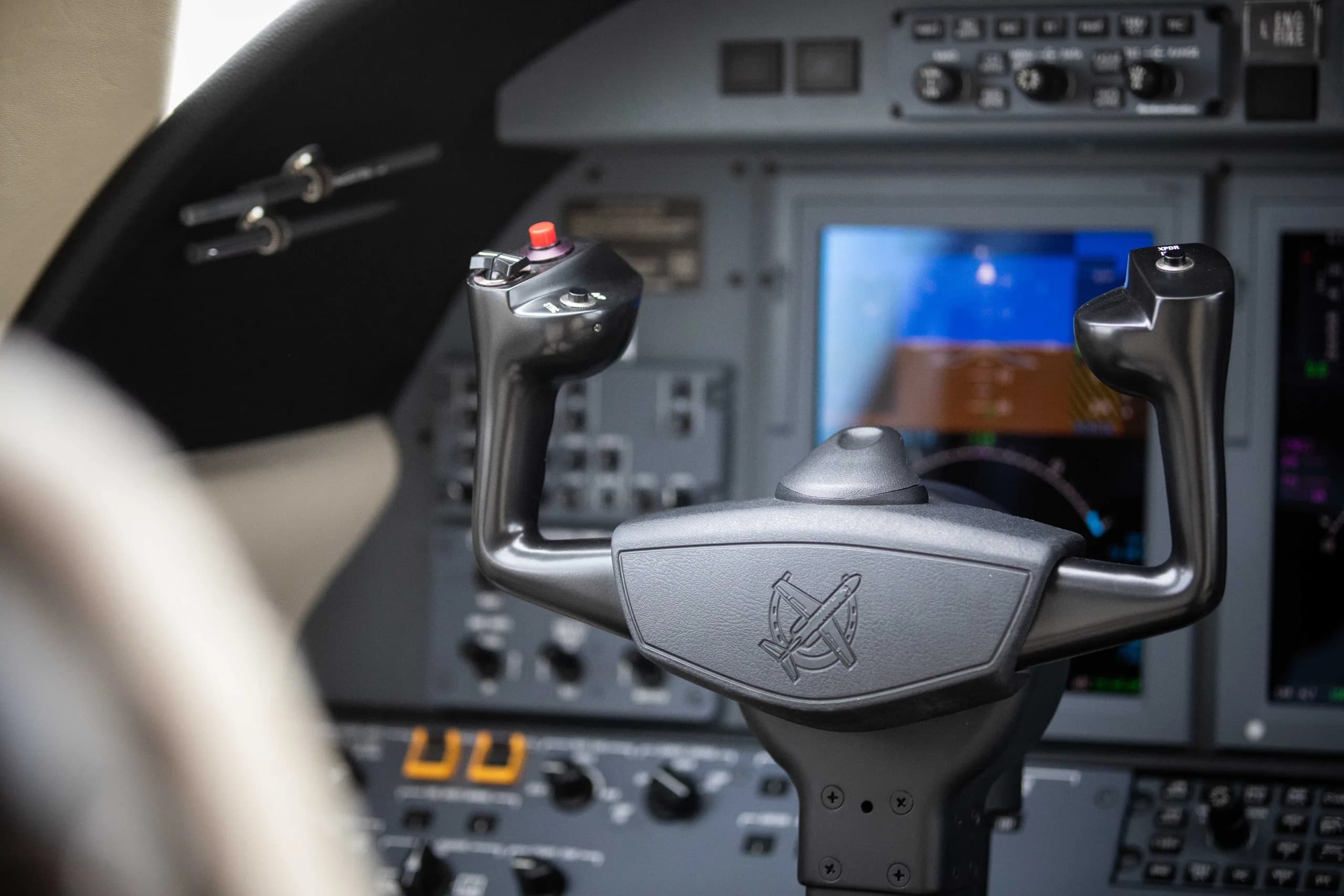 A close-up of an aircraft's control yoke with a red button and digital screens in the background.
