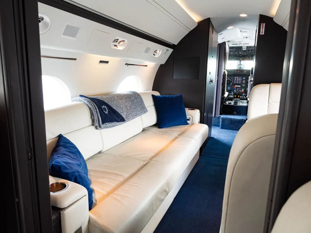 View of the aircraft interior with a white leather couch and blue pillows.