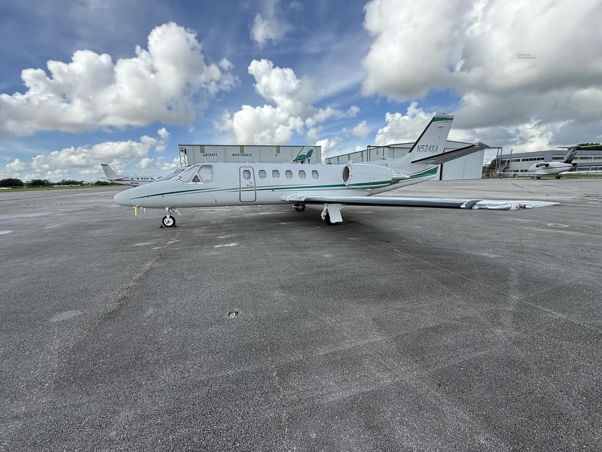 A white business jet N524XA with green stripes, parked on an airport tarmac with hangars and other aircraft in the background.