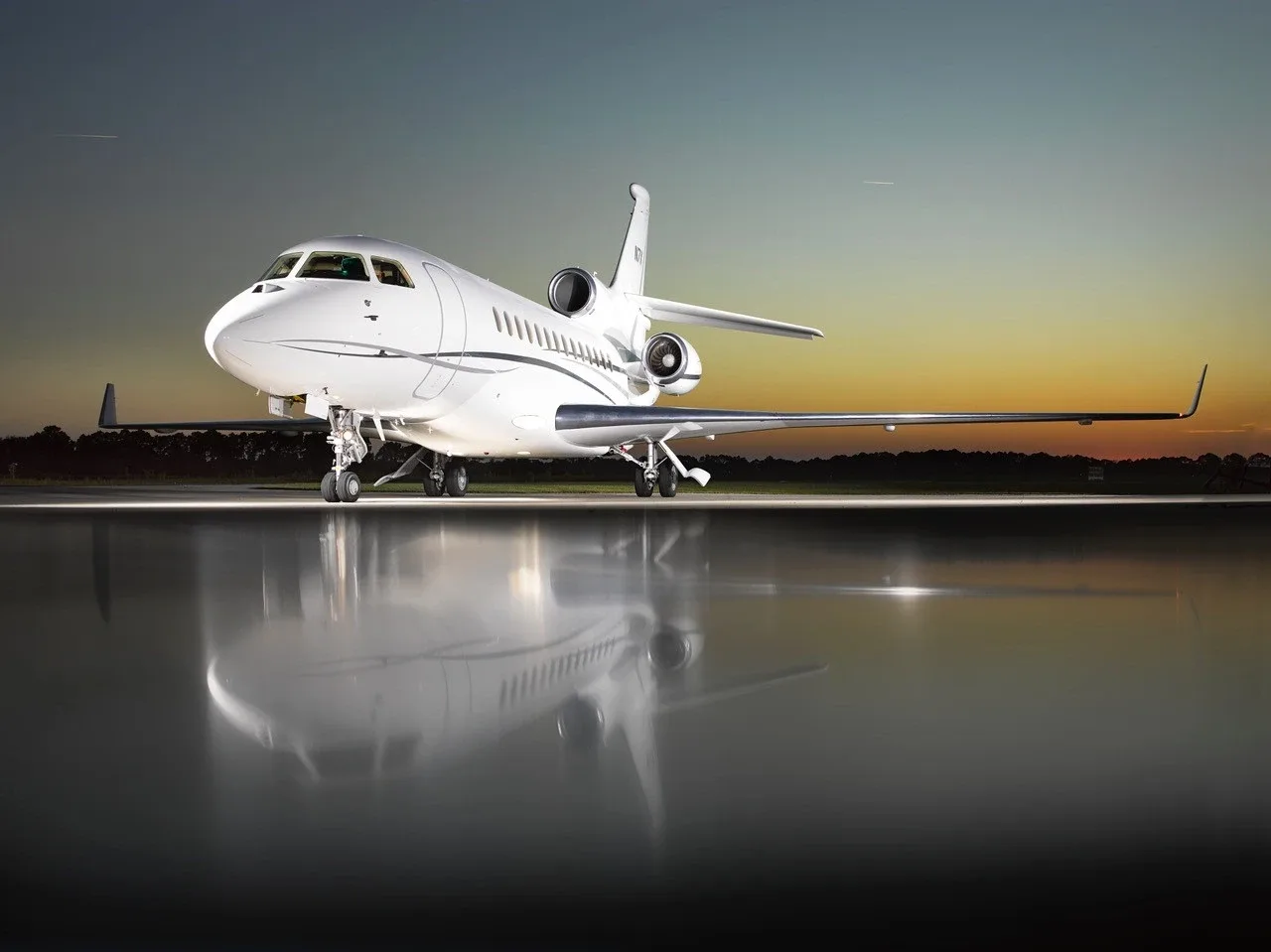 A white Dassault Falcon 7X business jet, registration N7X, parked on a wet tarmac at sunset, reflecting in the water.