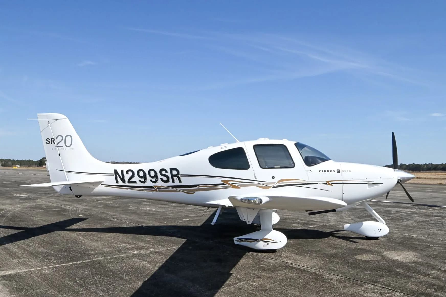 A white Cirrus SR20 aircraft with black and gold accent stripes, registration N299SR, parked on an airfield under a blue sky.