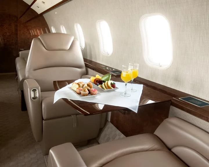 A close-up view inside a private jet cabin shows a table set with pastries, fruit, and two glasses of orange juice next to a beige leather seat.