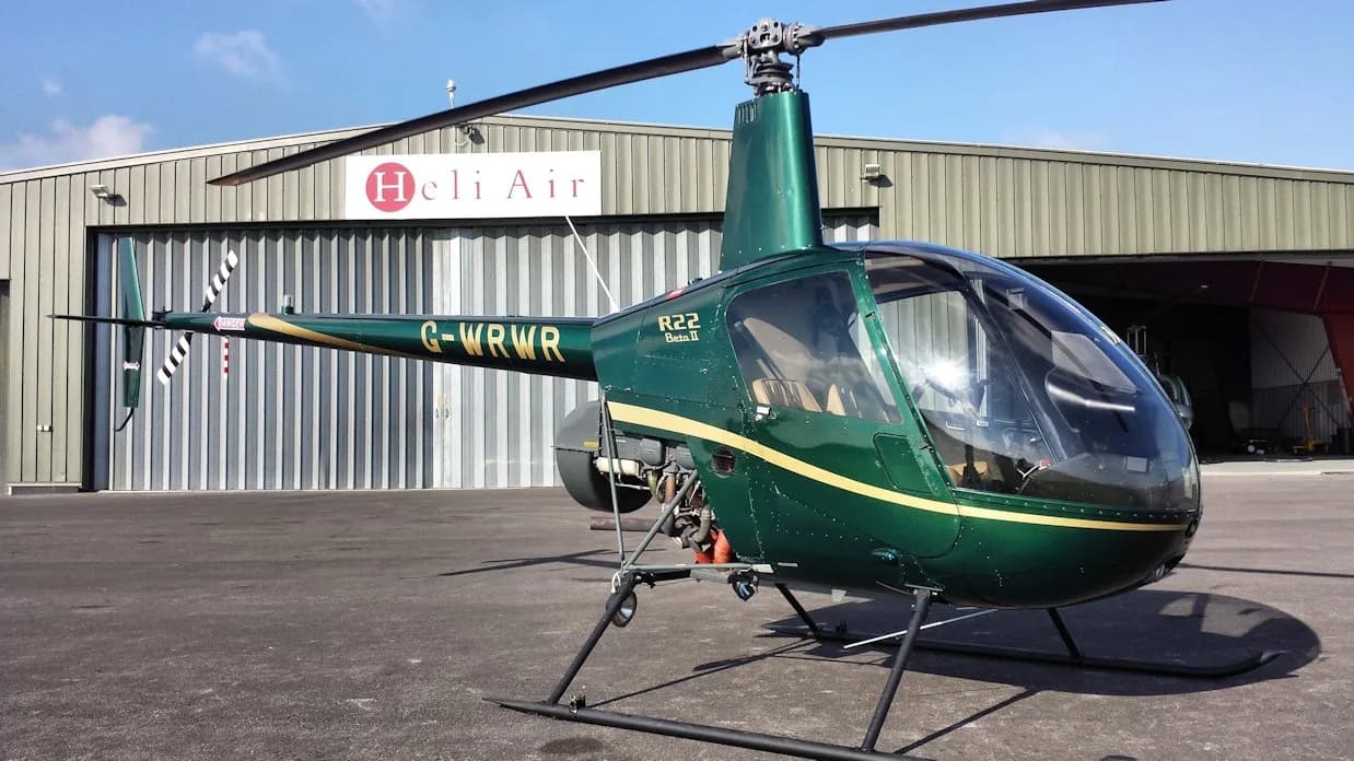 A dark green Robinson R22 Beta II helicopter (G-WRWR) with a gold stripe parked on a tarmac in front of a hangar.