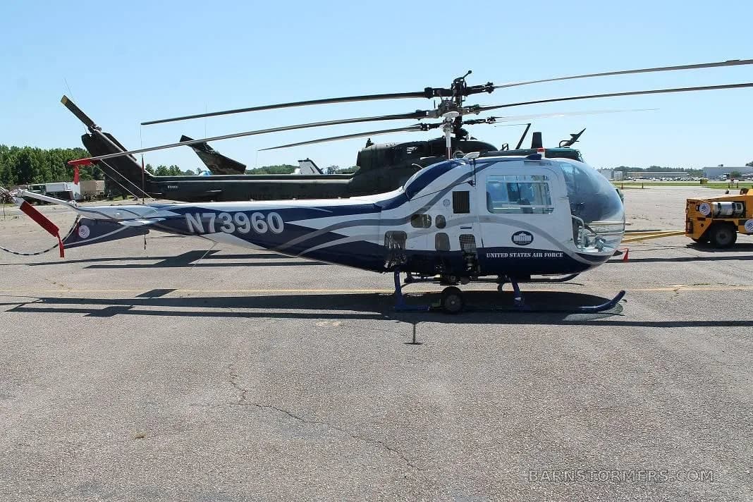 A side view of a white and blue helicopter with 'UNITED STATES AIR FORCE' text and registration N73960, parked on an airfield next to a larger dark helicopter.