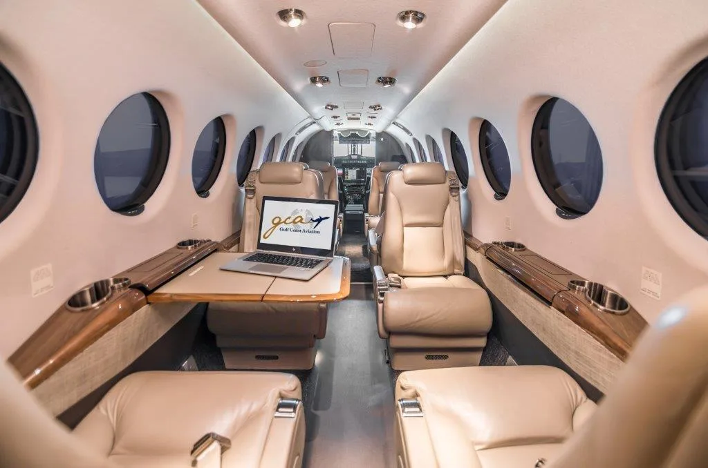 The interior cabin of a private jet with light brown leather seats and a laptop displaying a logo on a table.