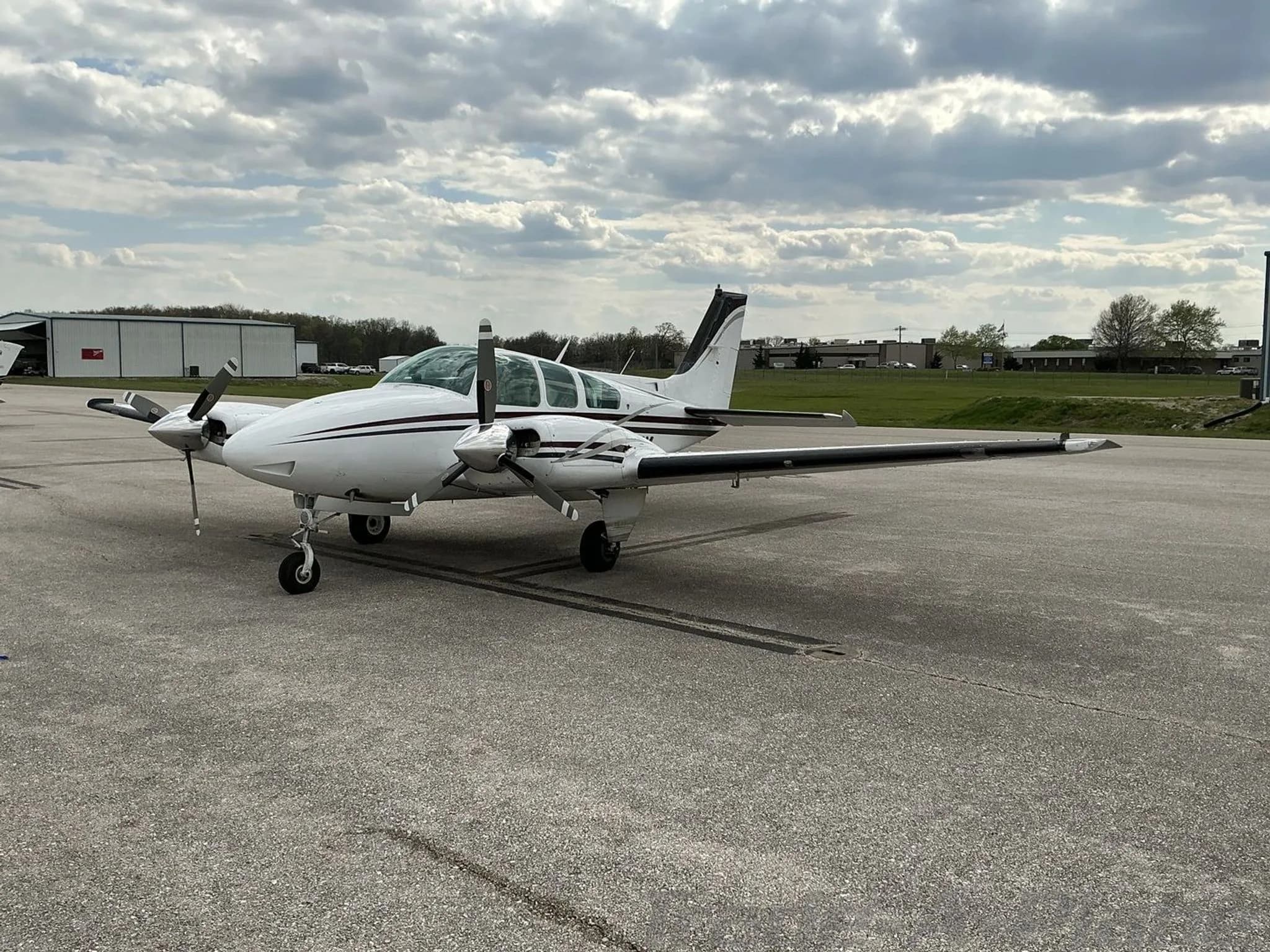 A white twin-engine propeller aircraft with black and maroon stripes parked on an asphalt tarmac under a cloudy sky.
