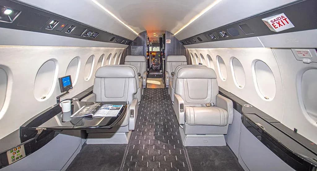 The spacious interior cabin of the Dassault Falcon 900EX, featuring light-colored leather seats and a table with a magazine.