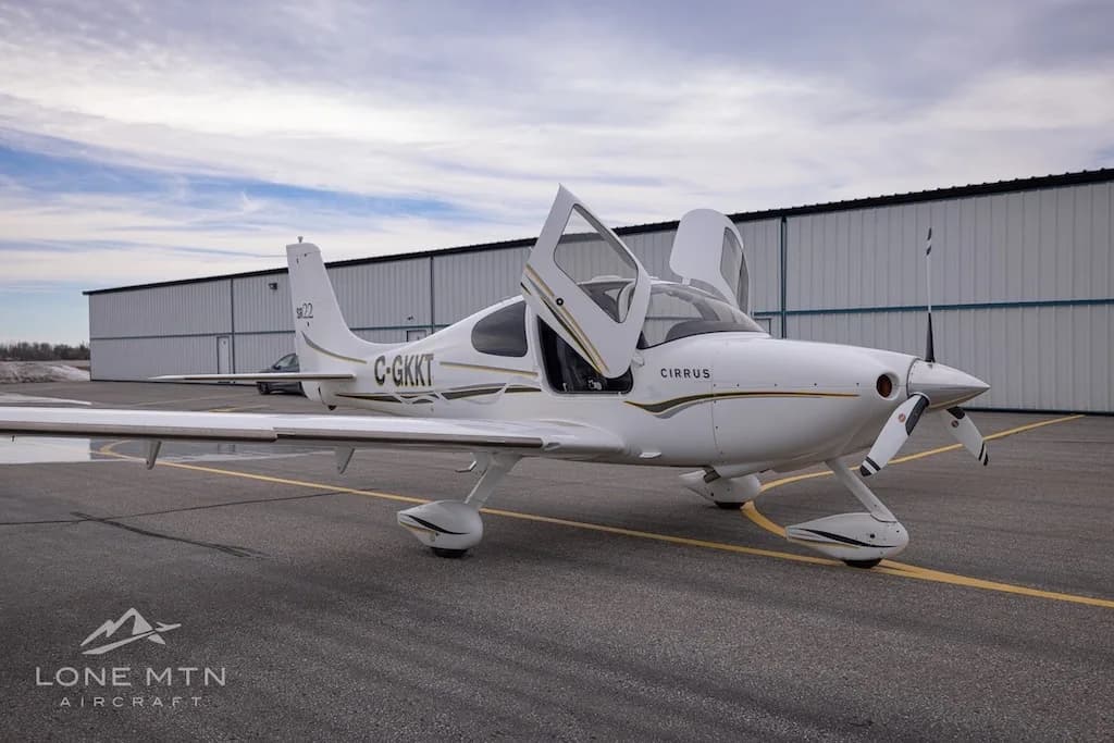 A front three-quarter view of a white Cirrus SR22 aircraft with registration C-GKKT, doors open, on an airfield.
