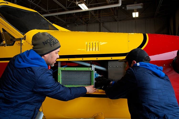 Two mechanics working on the engine of a yellow and red floatplane inside a hangar.
