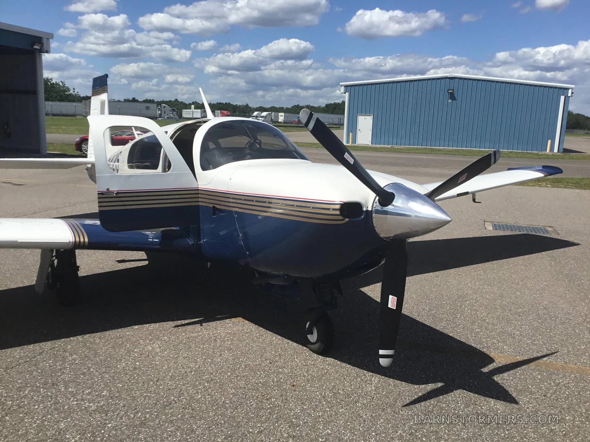 A white and blue single-engine propeller aircraft with its door open is parked on an asphalt ramp in front of a blue hangar.