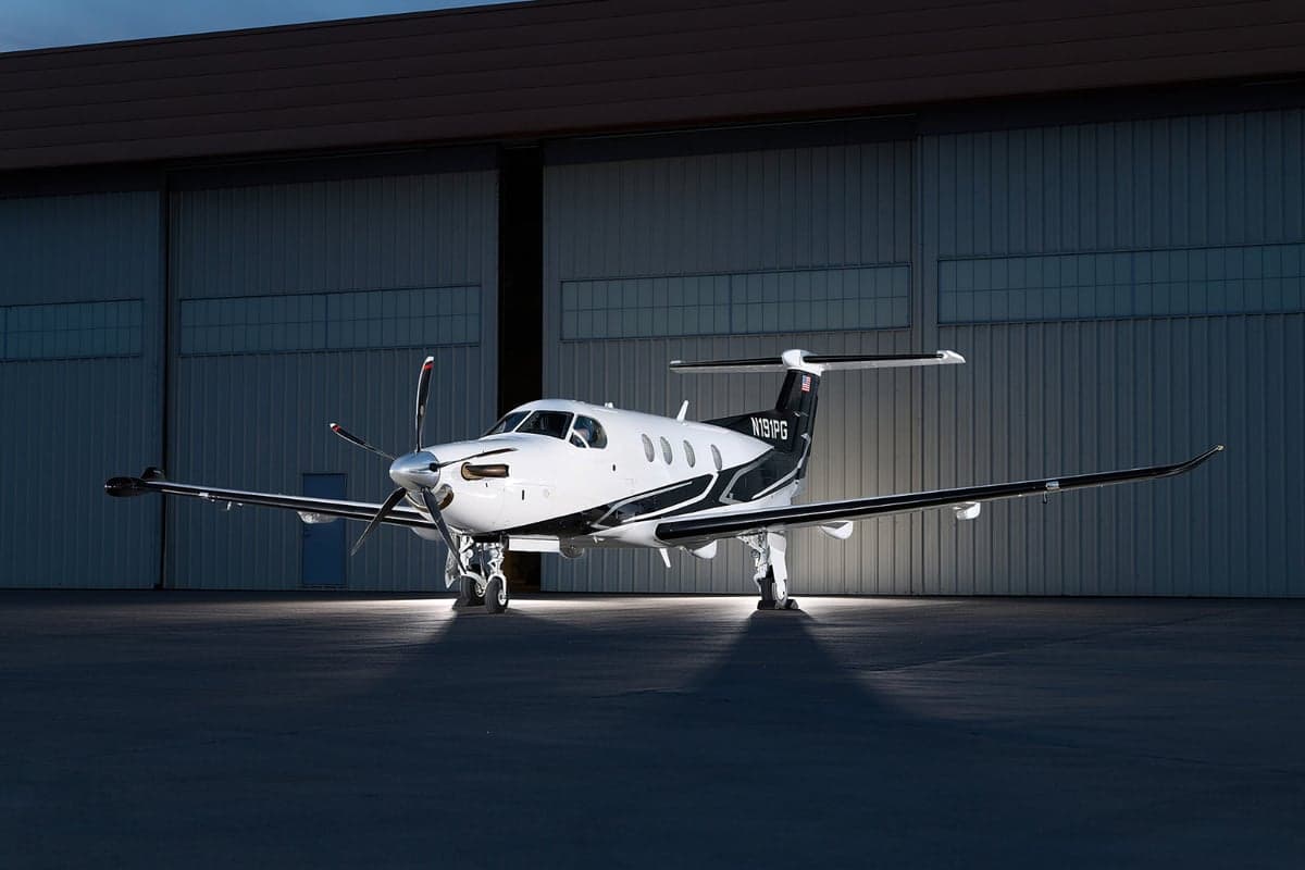 A white and black Pilatus PC-12 aircraft with tail number N191PG is parked in front of a hangar at dusk, dramatically lit from below.