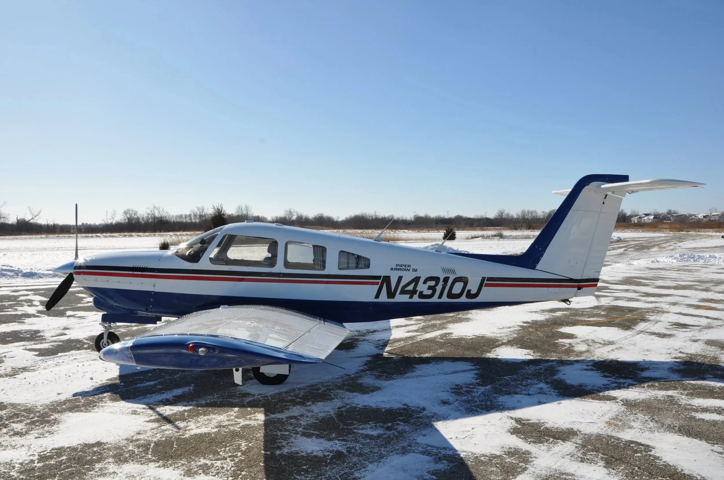 A full side view of the white and blue Piper Arrow IV aircraft with registration N4310J parked on a snowy tarmac.