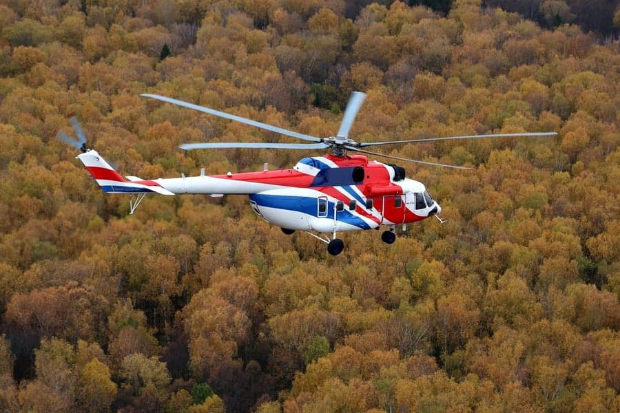A red, white, and blue Mil Mi-8 helicopter in flight over a forest.