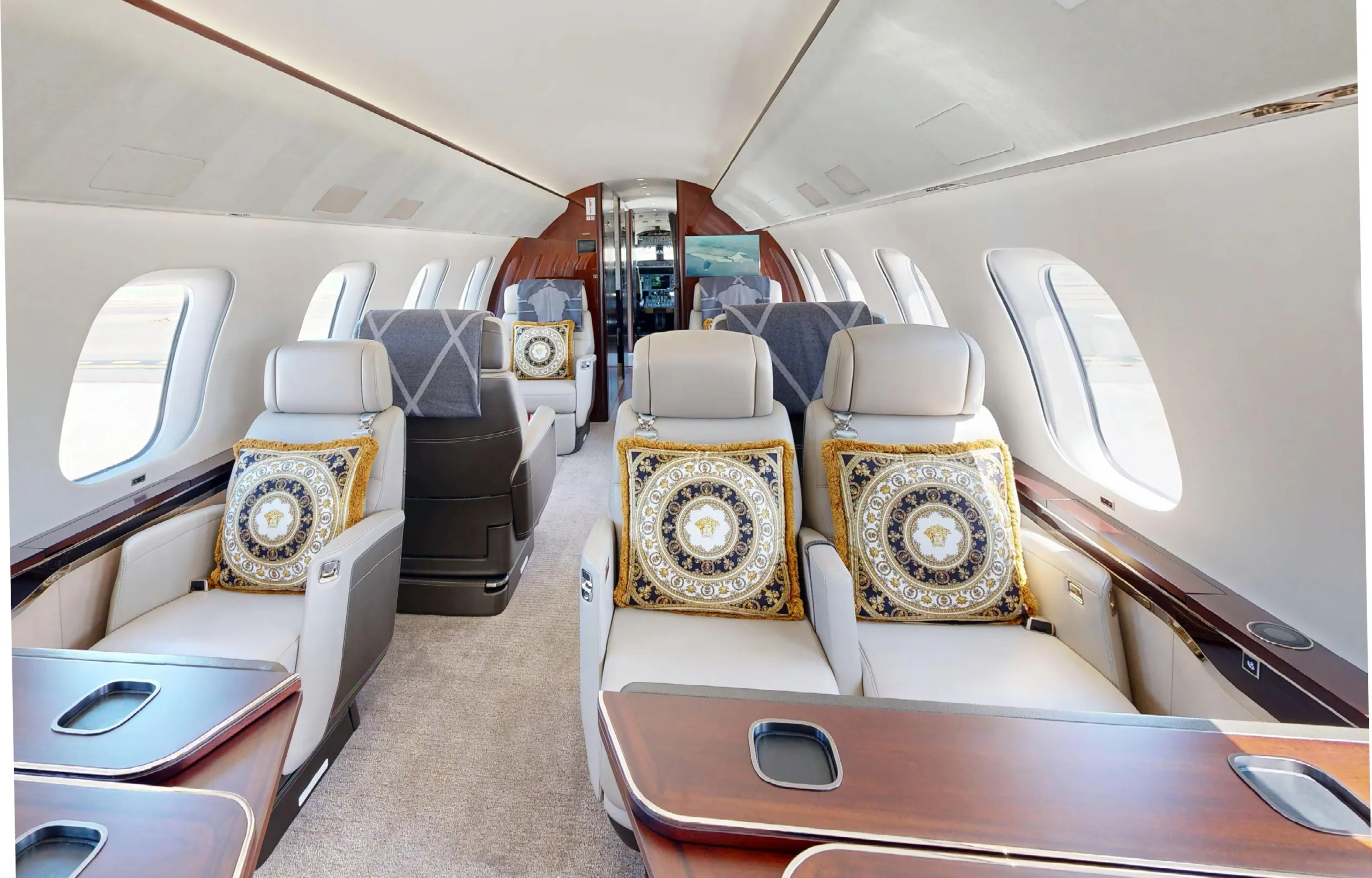 The luxurious cabin interior of a private jet, featuring light-colored seats with ornate gold and black Versace pillows and dark wood tables.