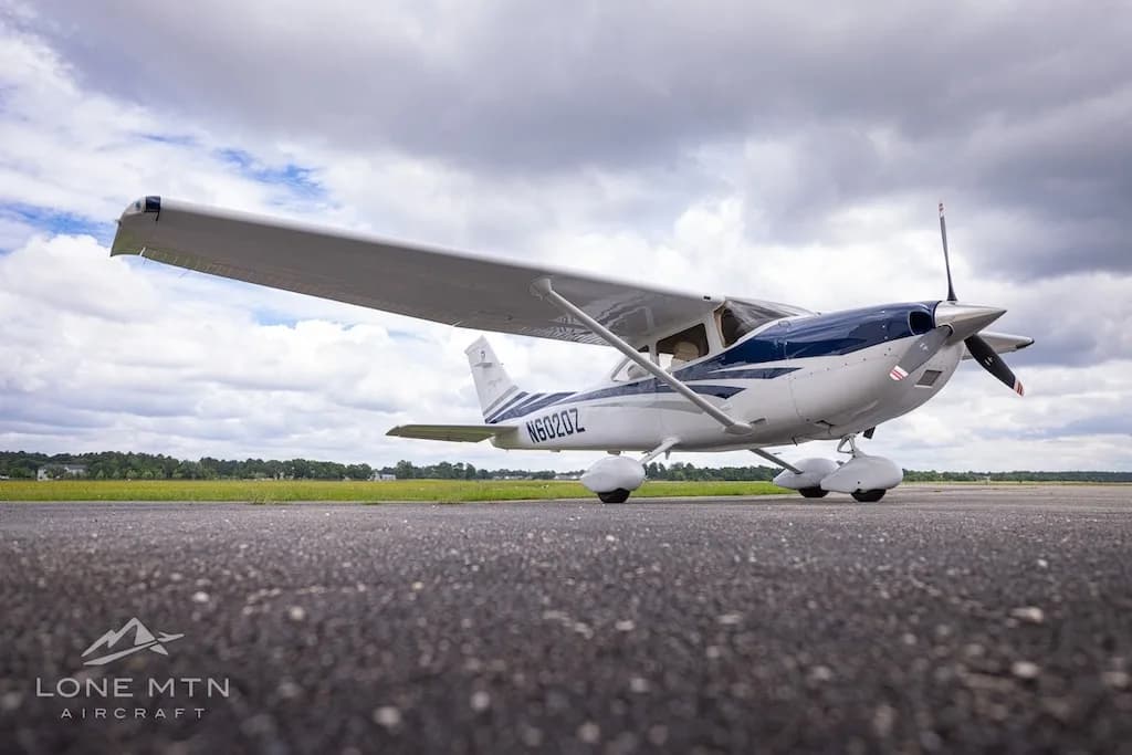 A low-angle front-right view of a white and blue Cessna Skylane TC (N6020Z) on an airfield under a cloudy sky.