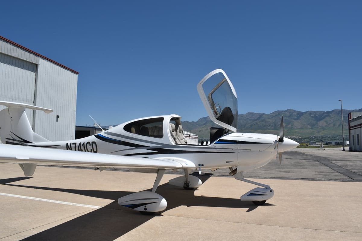 A white Diamond DA40 aircraft (N741CD) with blue and grey stripes, parked on an airfield with its canopy open and mountains in the background.