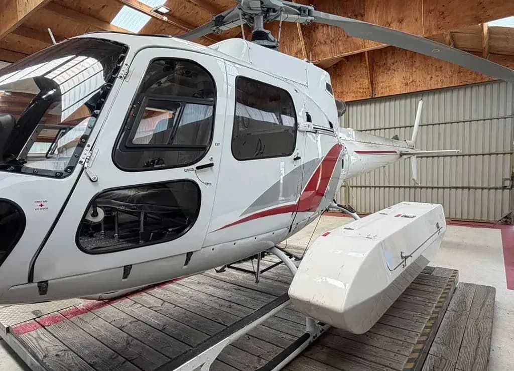 A white helicopter with red and grey stripes, parked on a wooden platform inside a hangar, viewed from the side, showing an external cargo pod.