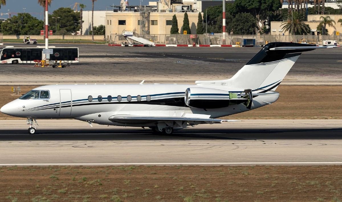 A white business jet with black and blue stripes on its fuselage and tail, seen from the side on an airport tarmac.