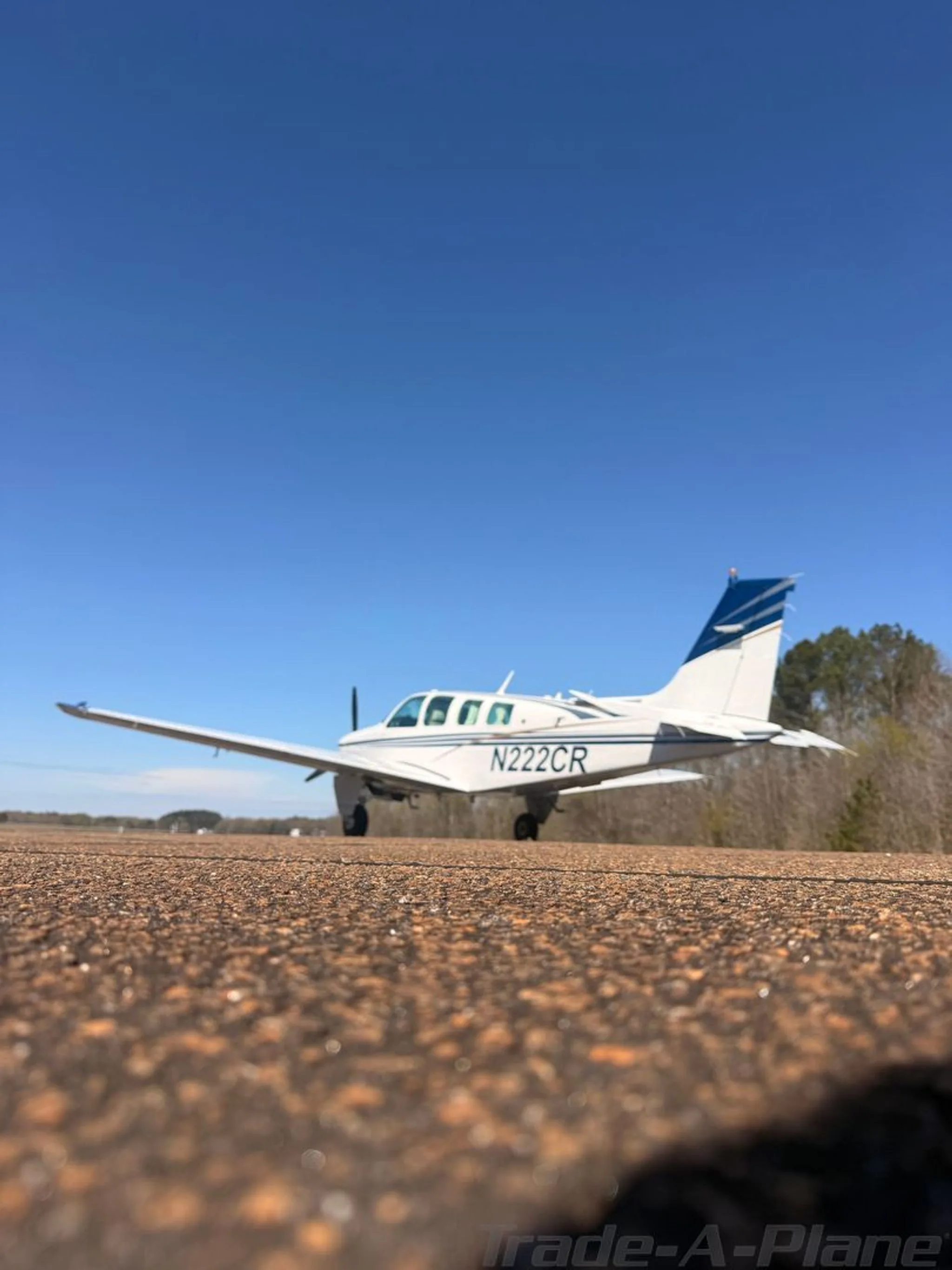 A low-angle view of a white Beechcraft Bonanza N222CR with blue and gold stripes on an asphalt runway.