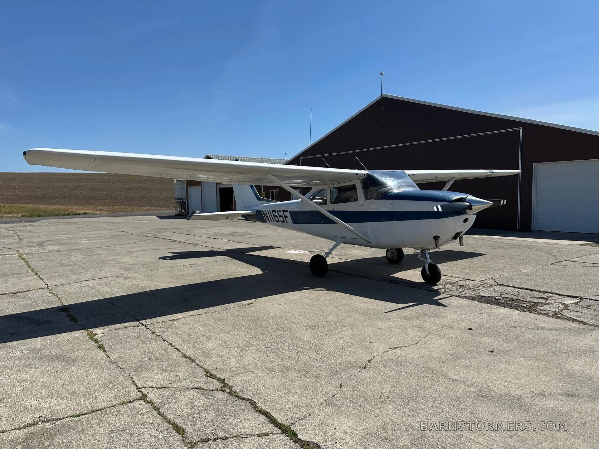 A white and blue Cessna N1165F aircraft is parked on a concrete apron in front of a red hangar on a sunny day.