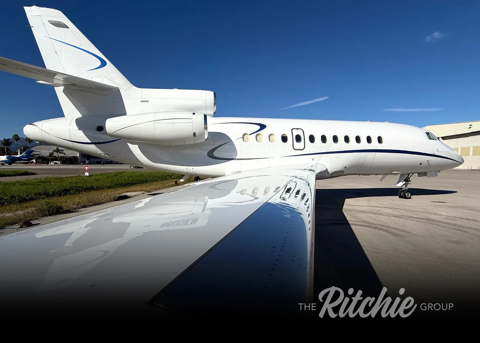 A white business jet with blue accents, viewed from the rear-side showing the wing and engines, parked on an airfield under a clear blue sky.