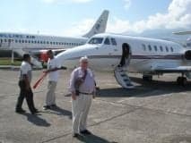 A white aircraft is parked on the tarmac with people standing nearby.