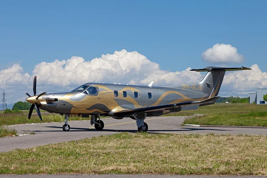 A silver and gold Pilatus PC-12 aircraft (OH-RJD) with a unique paint scheme, parked on an airfield under a partly cloudy blue sky.