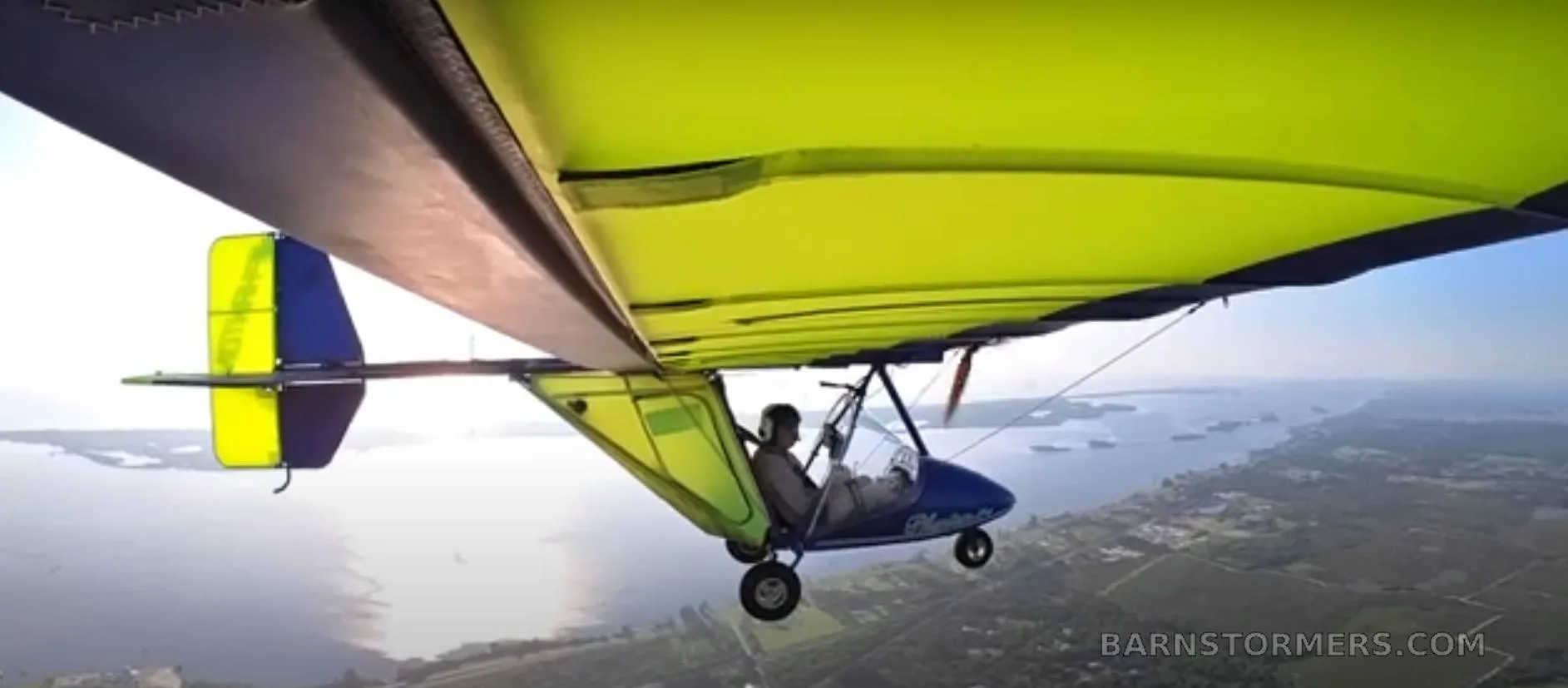 A blue and yellow ultralight aircraft with a bright green wing flying over water and land, viewed from below.
