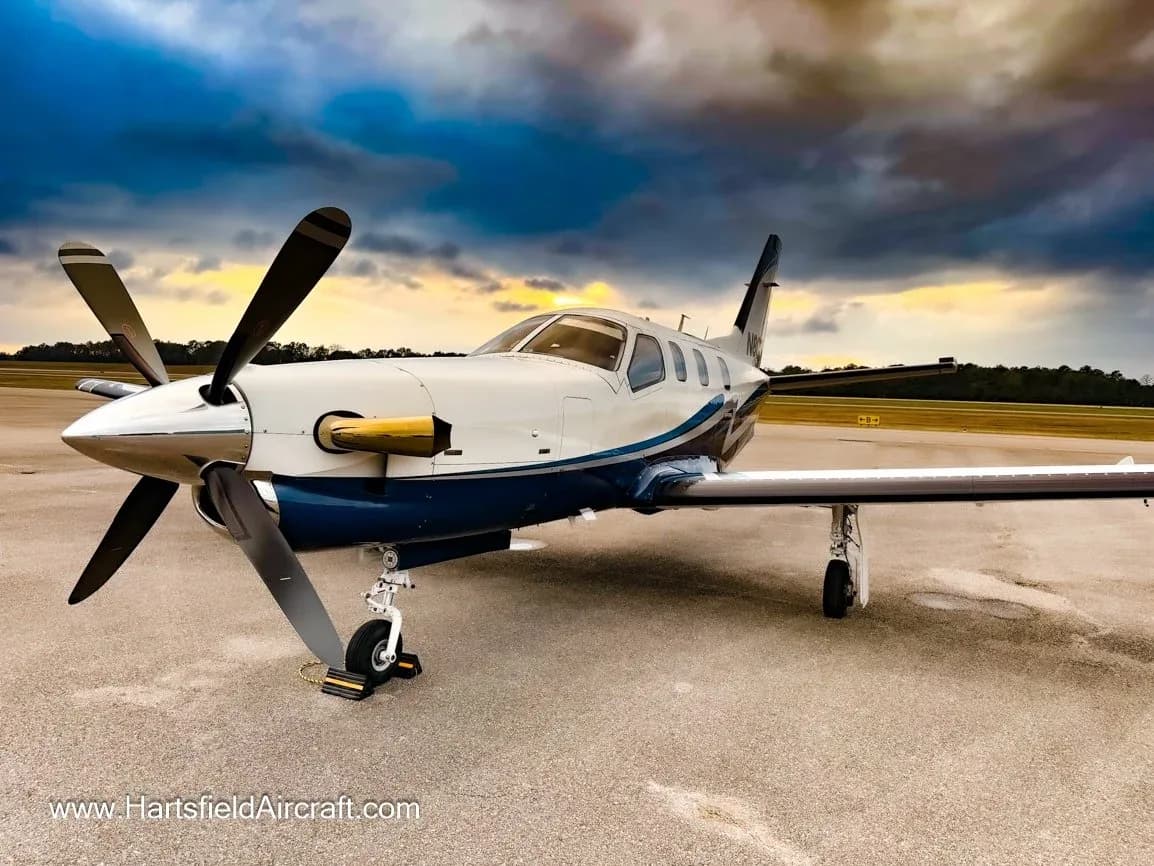 A white and blue single-engine turboprop aircraft with a four-blade propeller parked on an airfield under a dramatic sunset sky.