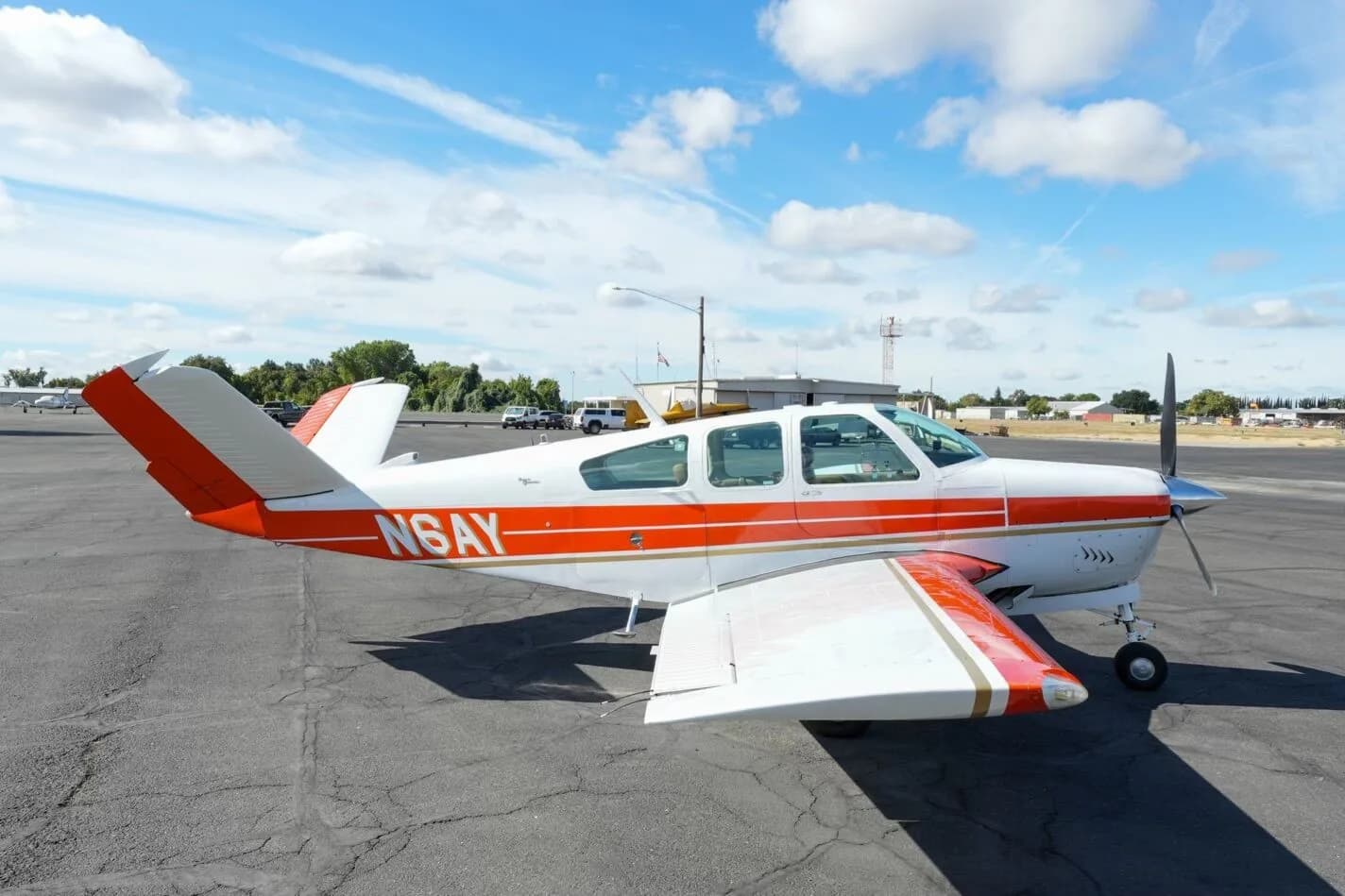 A three-quarter view of the white and orange Beechcraft Bonanza N6AY on the tarmac, facing right.