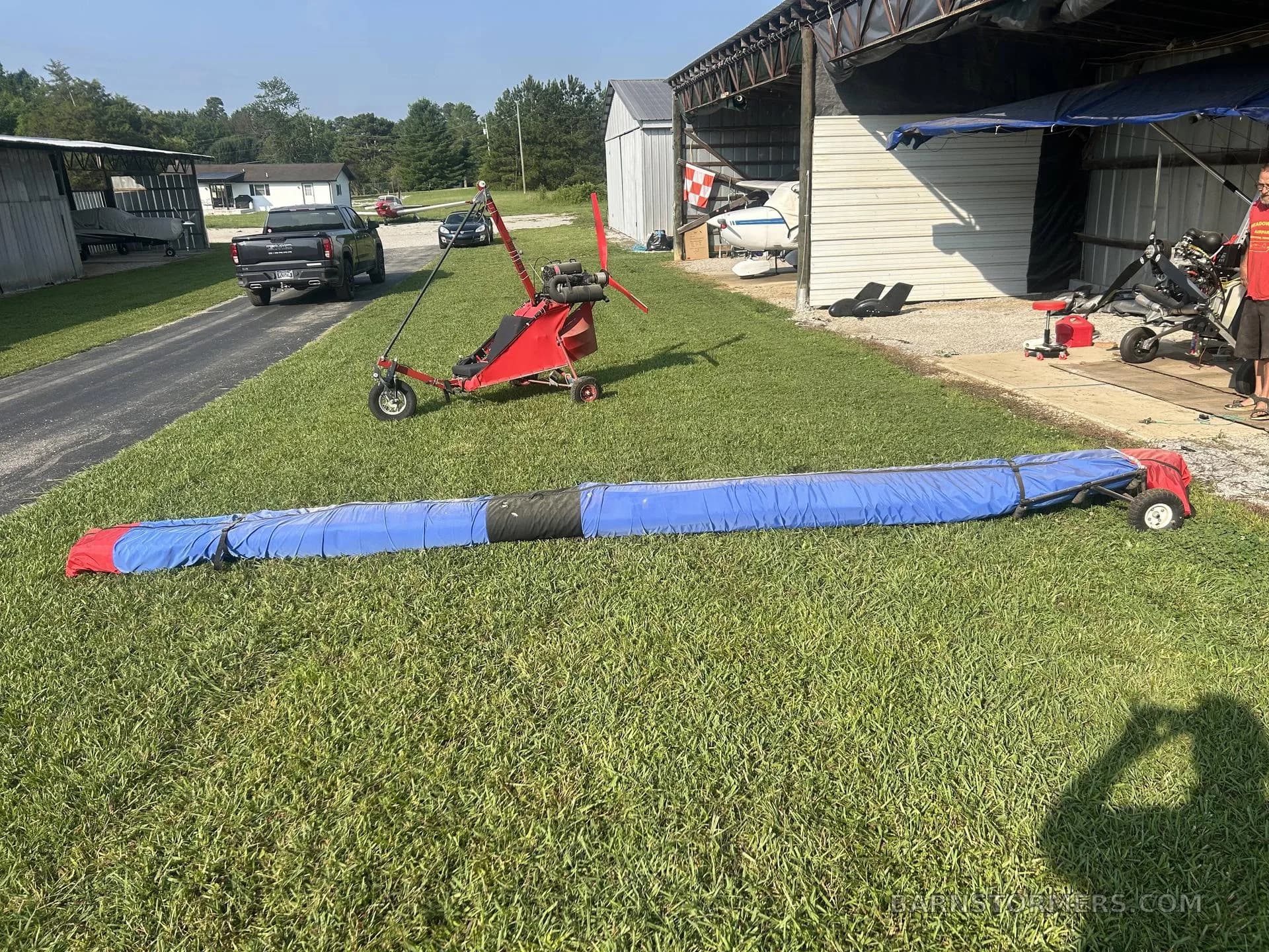 Several ultralight aircraft, including a red one and a blue covered wing, parked on a grassy field near hangars.