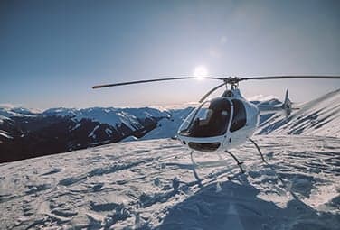 A white helicopter parked on a snowy mountain top with a mountain range in the background.