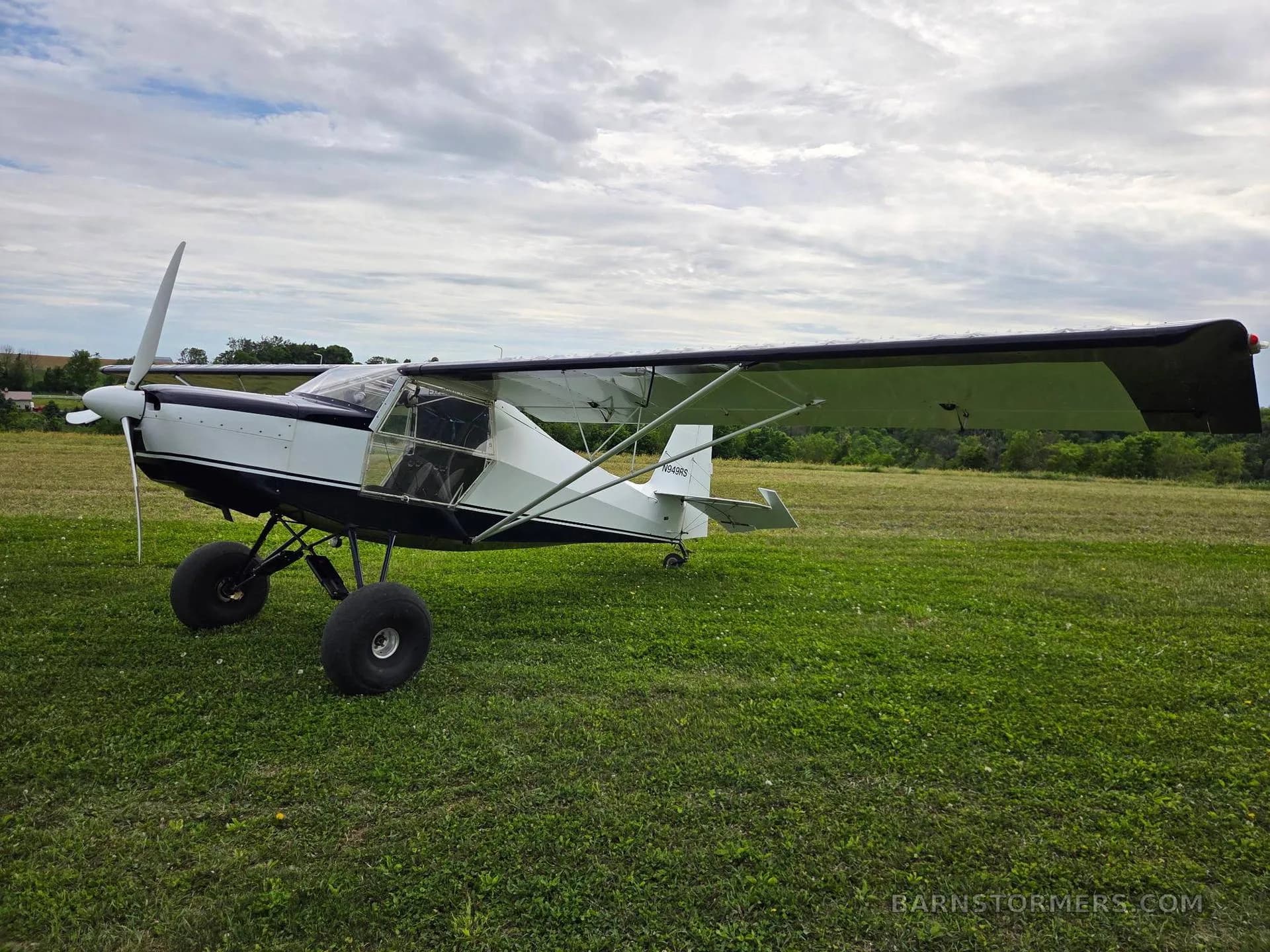 A white and black single-engine aircraft with large tires is parked on a grassy field under a cloudy sky.