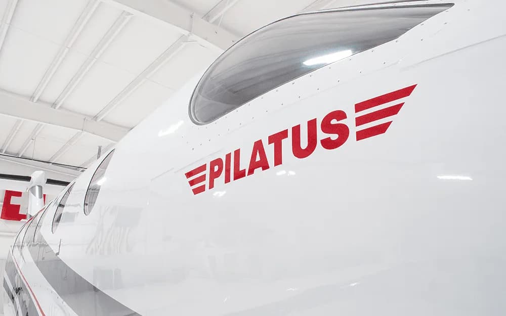 A close-up view of the white fuselage of a Pilatus PC-12 showing the red "PILATUS" logo and cockpit window.