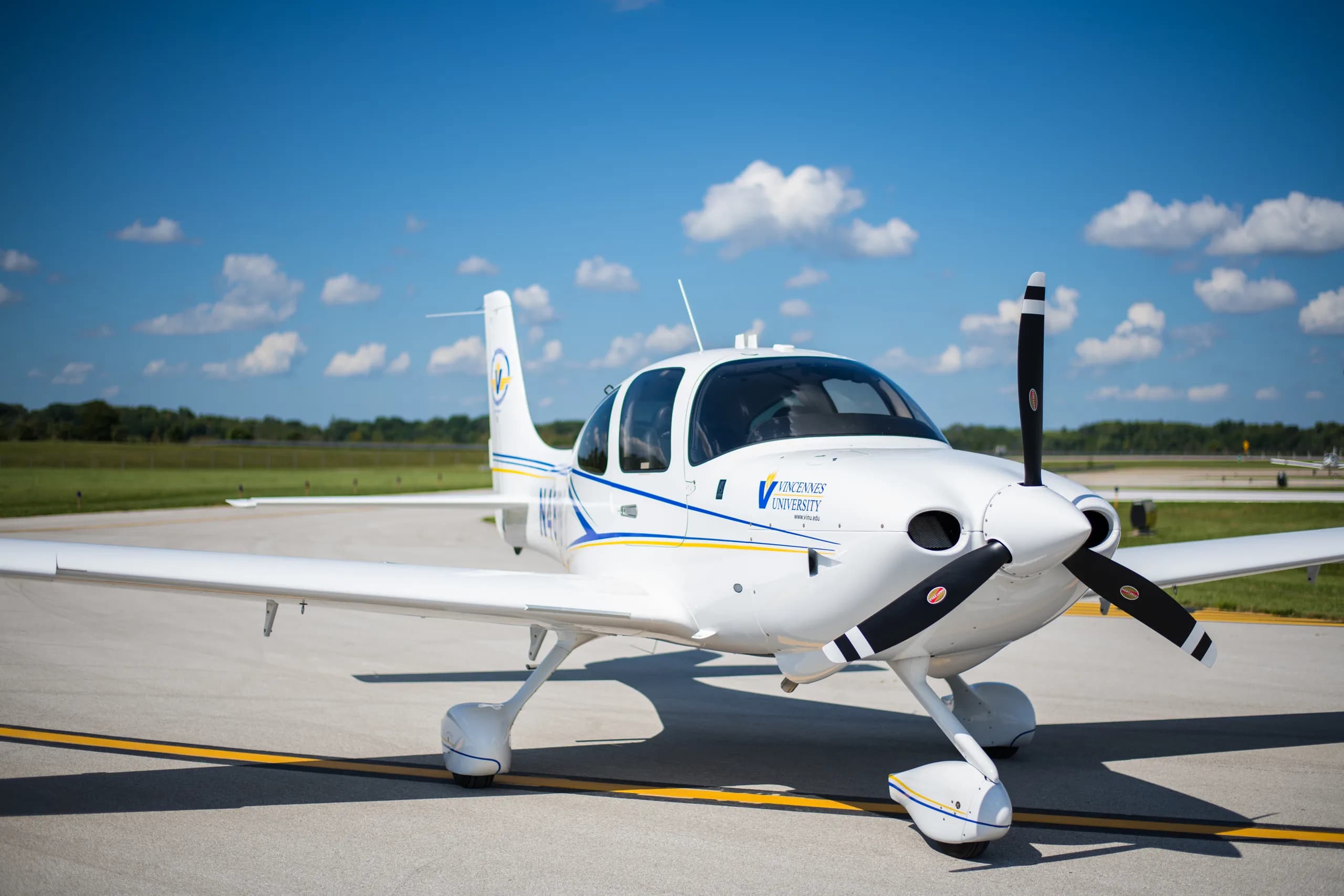 A white single-engine propeller aircraft with blue and yellow stripes and "Vincennes University" branding, parked on an asphalt runway under a blue sky with clouds.