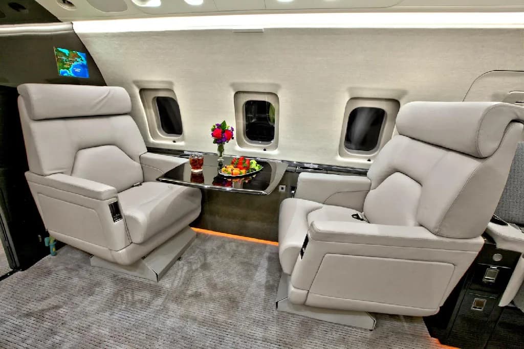 Two plush light gray individual seats in the private jet cabin, with a small table between them holding drinks and fruit.